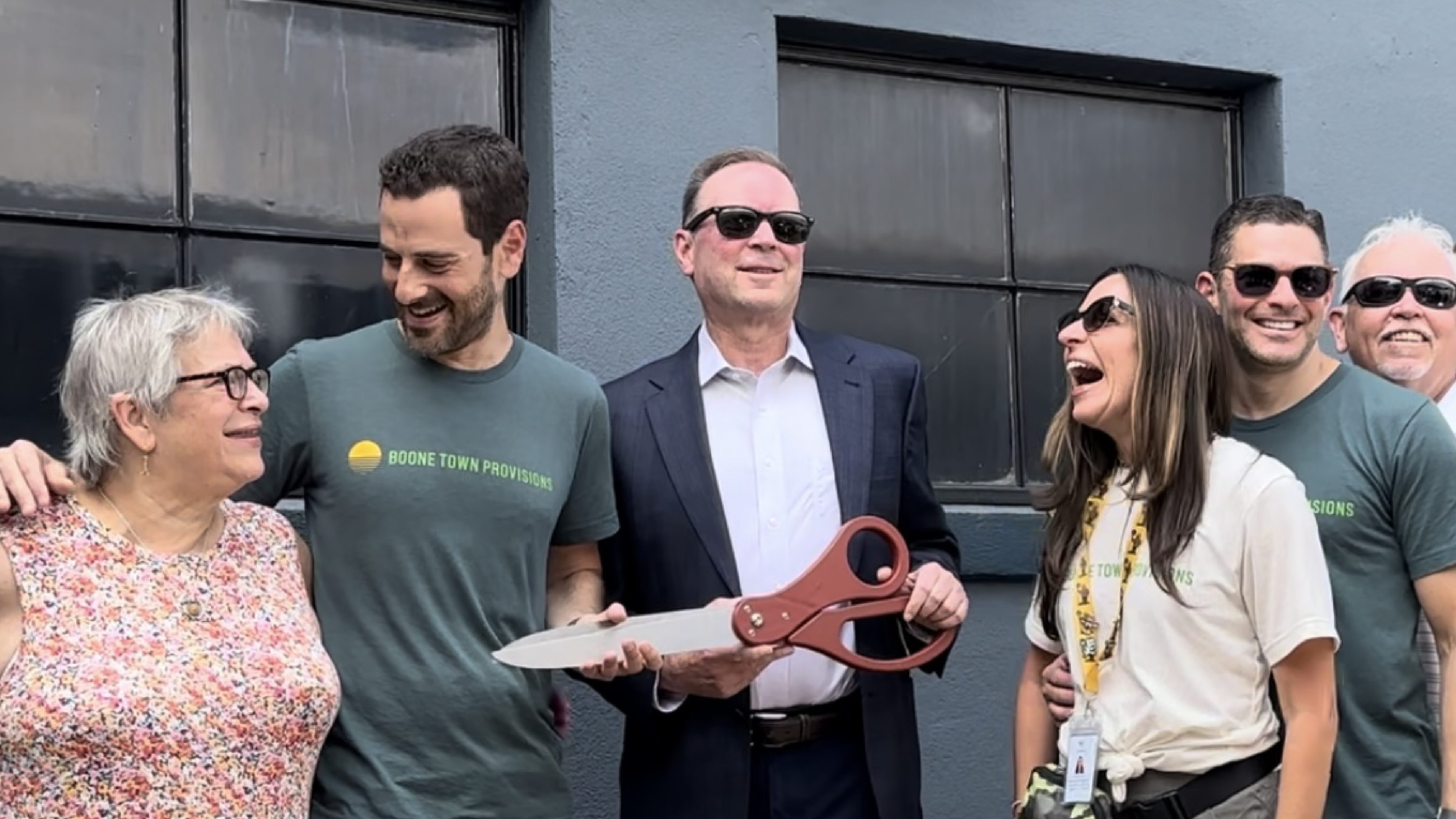 Boone Town Provisions team at their grand opening, gathered together with large scissors for the ribbon-cutting ceremony, sharing smiles and laughter.