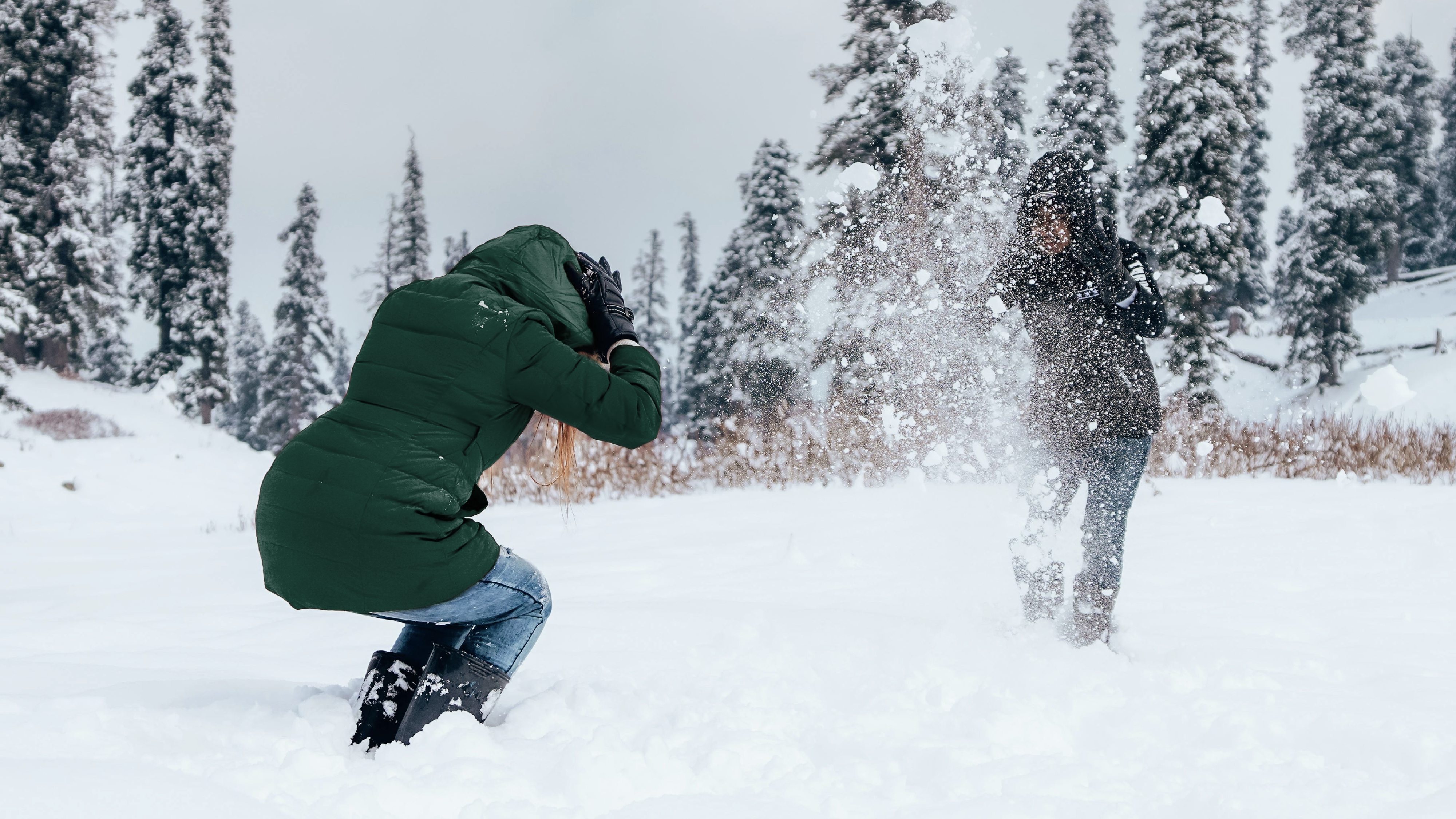 Two people having a playful snowball fight in a snowy forest, with snow flying in the air as one person ducks.